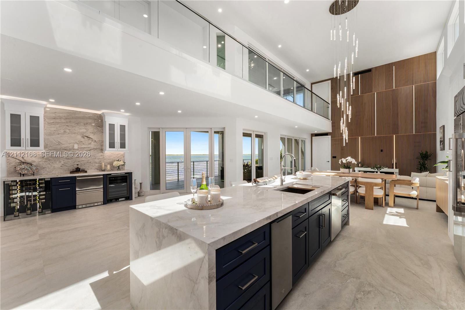 Double-height ceilings with custom wood-panel wall feature, oversized kitchen island with stone waterfall countertop, integrated floor-to-ceiling glass panel balcony railing, contemporary tiered pendant lighting, and multiple sets of glass exterior doors