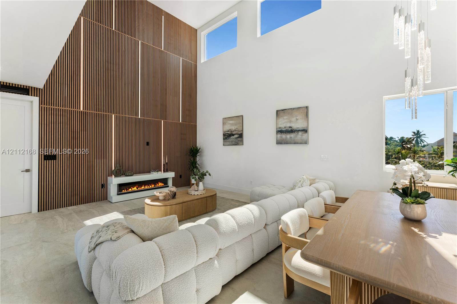 Double-height ceilings with clerestory windows, vertical slat wood-finish accent wall with integrated lighting, built-in linear electric fireplace, stone-finish flooring, and multi-light hanging chandelier
