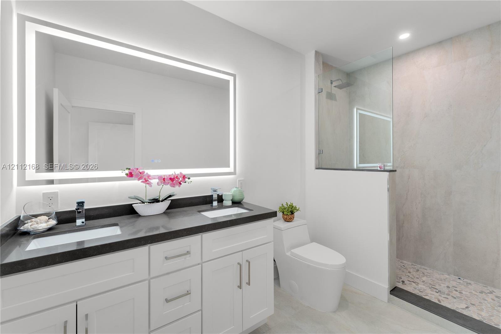 Double vanity with dark countertops, integrated rectangular sinks, wall-to-wall LED backlit mirror, walk-in glass shower with stone-style tile surround, and tile flooring