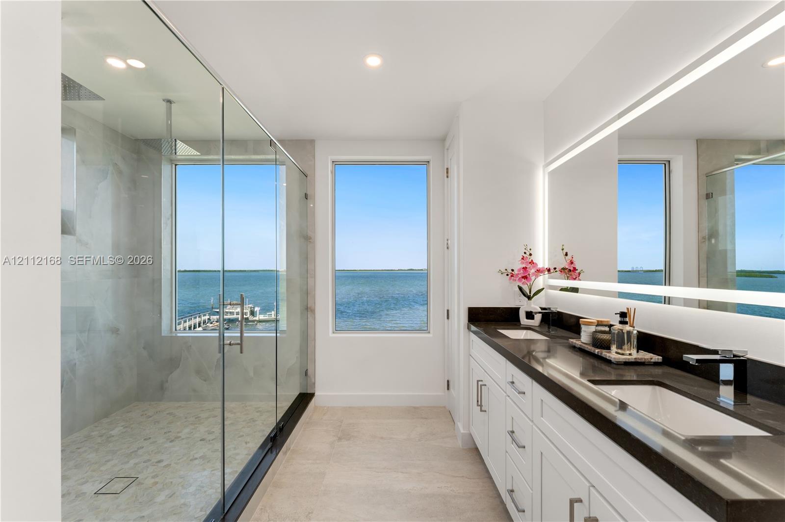 Dual-vanity bathroom featuring white cabinetry, dark stone countertops, a large frameless glass-enclosed shower with stone tile flooring, and a full-length illuminated vanity mirror