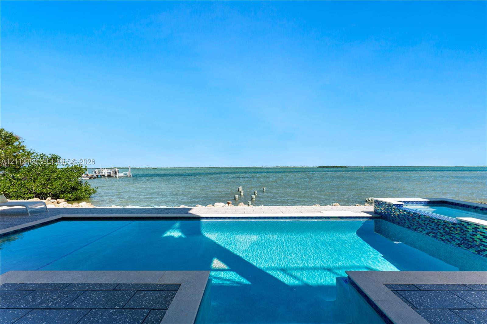 In-ground swimming pool featuring a tiled spillover spa, stone paver decking, and direct water frontage