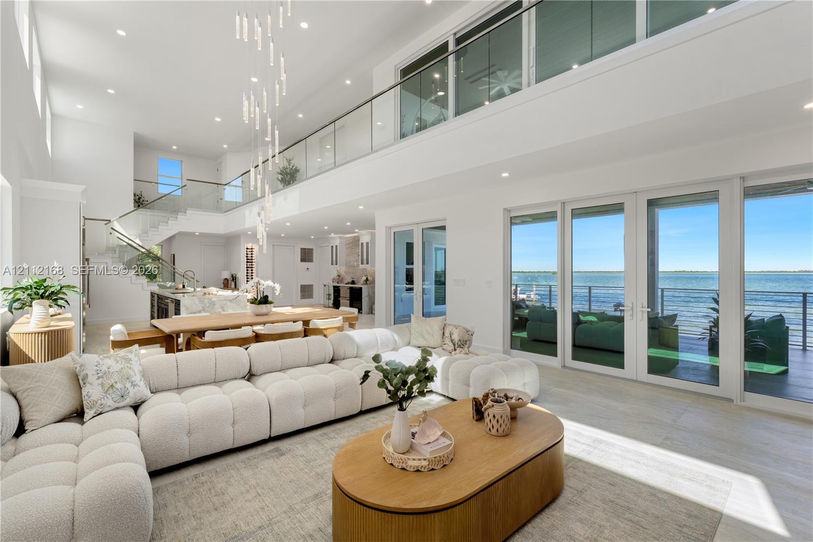 Expansive open-concept living area featuring double-height ceilings, a floor-to-ceiling glass-paneled mezzanine, multi-panel sliding glass doors, and a linear pendant light fixture