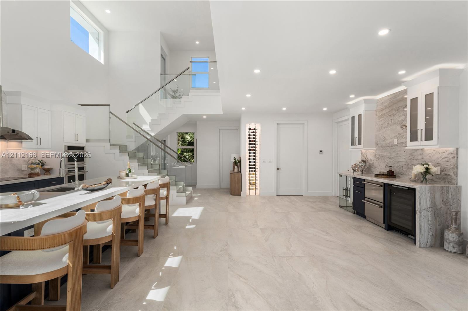 Open-concept kitchen featuring a large central island with waterfall-edge countertop, natural stone-style backsplash, integrated wine refrigeration, built-in vertical wine rack, and a glass-paneled staircase