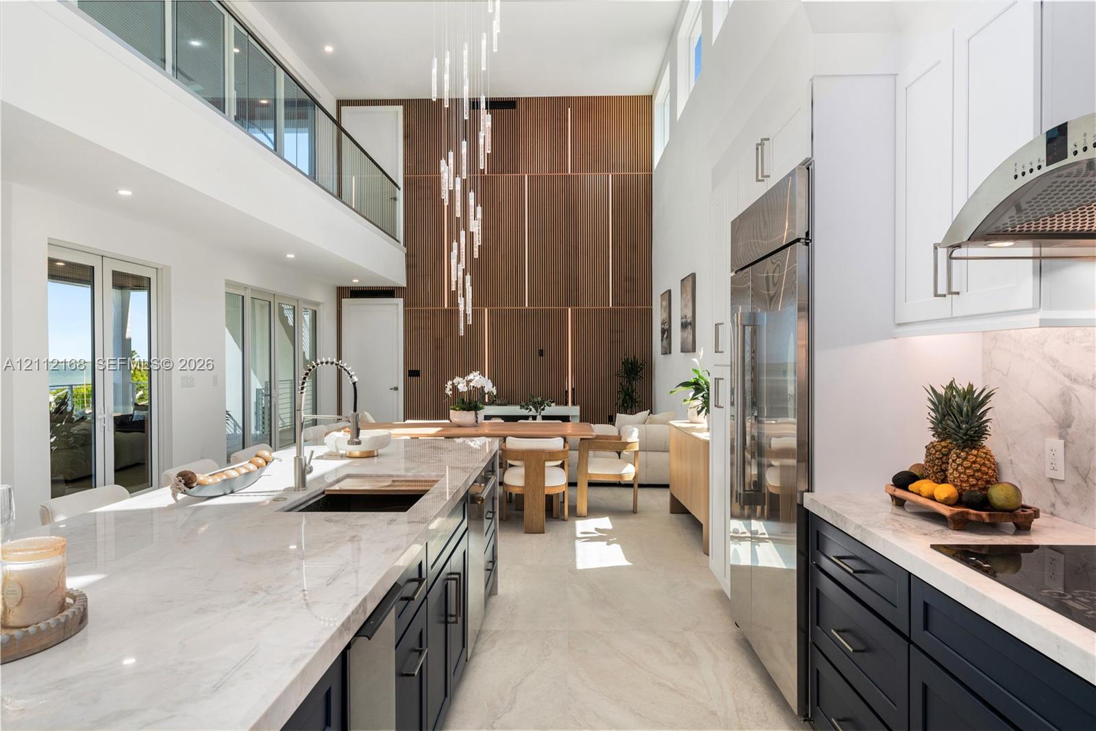 Double-height ceilings, floor-to-ceiling wood-panel accent wall, dual-tone cabinetry with stone-finish countertops, stainless steel integrated appliances, and glass-railing mezzanine