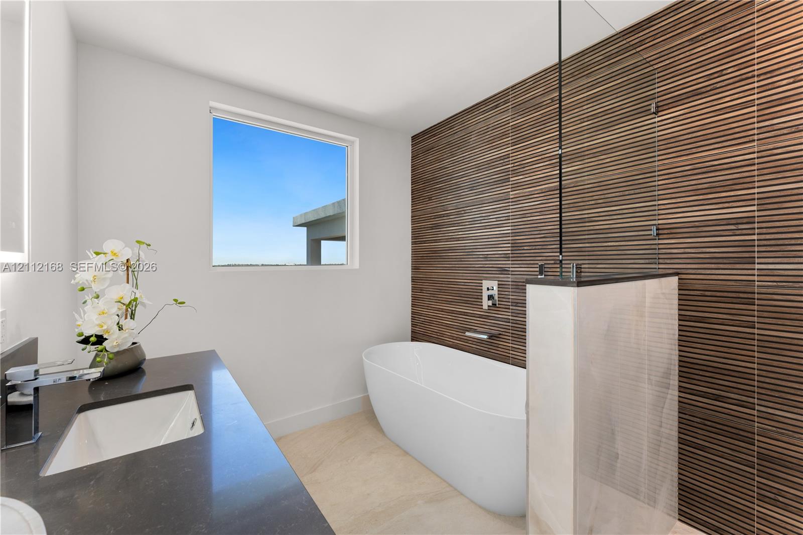 Freestanding soaking tub featuring a wall-mounted faucet, horizontal slatted wood-texture accent wall, glass partition, stone-finish vanity countertop, and rectangular undermount sink
