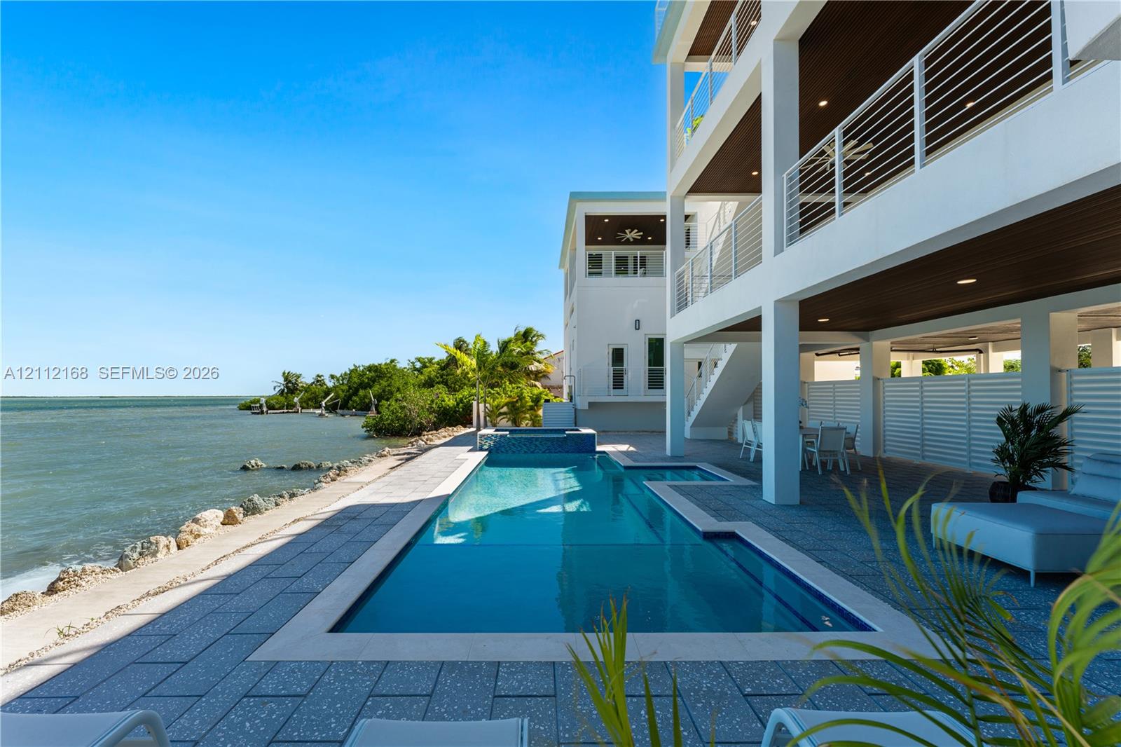 Waterfront exterior featuring a multi-level structure with recessed balconies, horizontal cable railings, a rectangular swimming pool with an integrated spa, and expansive stone-tiled patio decking
