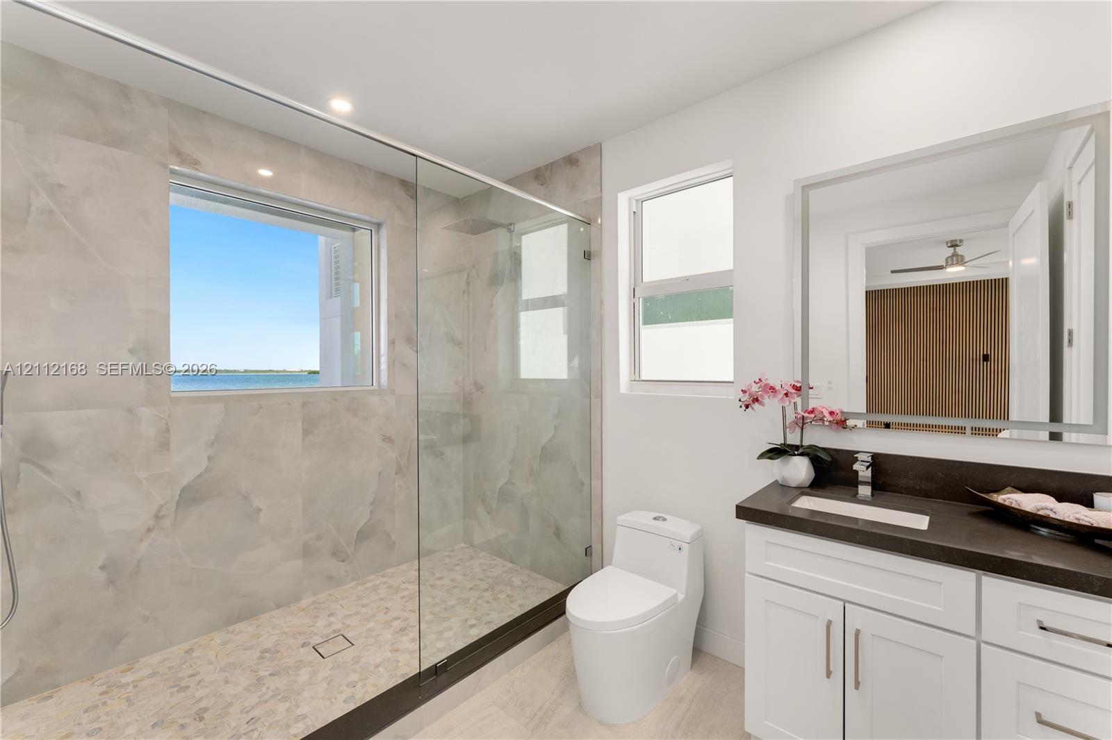 Bathroom featuring a walk-in shower with large-format wall tile and pebble-texture flooring, frameless glass enclosure, dual-pane window, and white cabinetry with a contrasting dark countertop