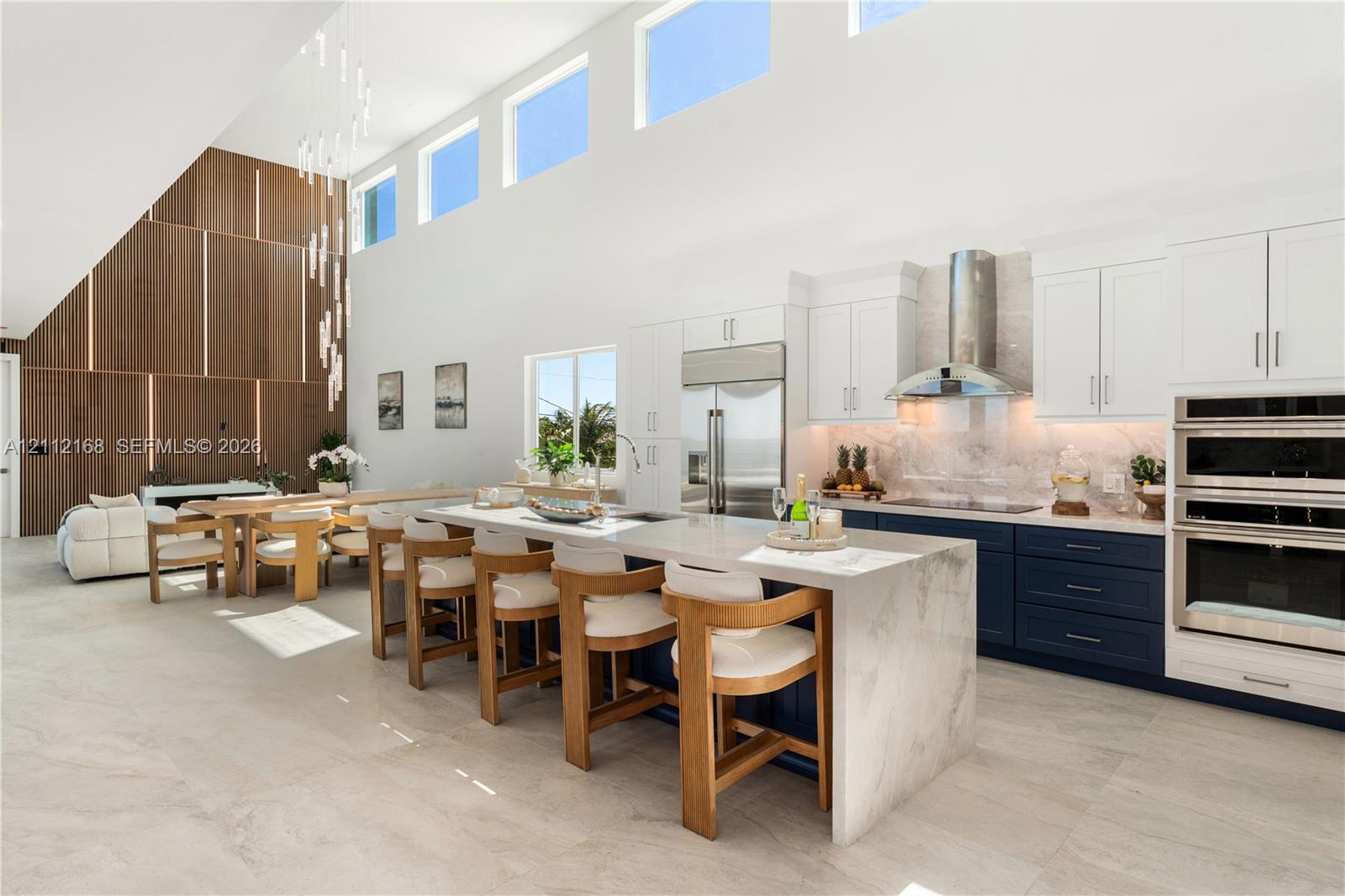 Open-concept kitchen featuring double-height ceilings, a large stone-topped waterfall island, custom two-toned cabinetry, built-in stainless steel appliances, and a floor-to-ceiling wood panel accent wall