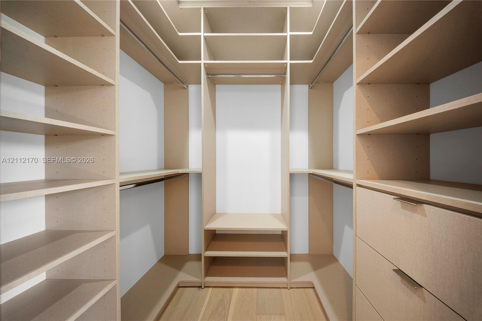 Custom walk-in closet featuring light-toned wooden built-in shelving, integrated hanging rods, three lower storage drawers with metallic pulls, and wood-finish flooring