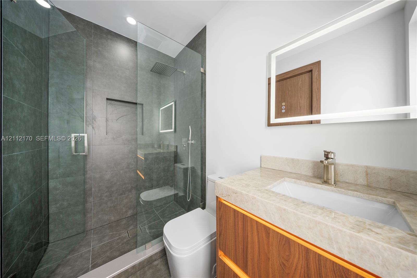 Bathroom featuring a glass-enclosed walk-in shower with large-format stone-finish tile and a built-in recessed niche, paired with a vanity featuring a stone countertop, integrated sink, and wood-finish cabinetry, complemented by a backlit vanity mirror