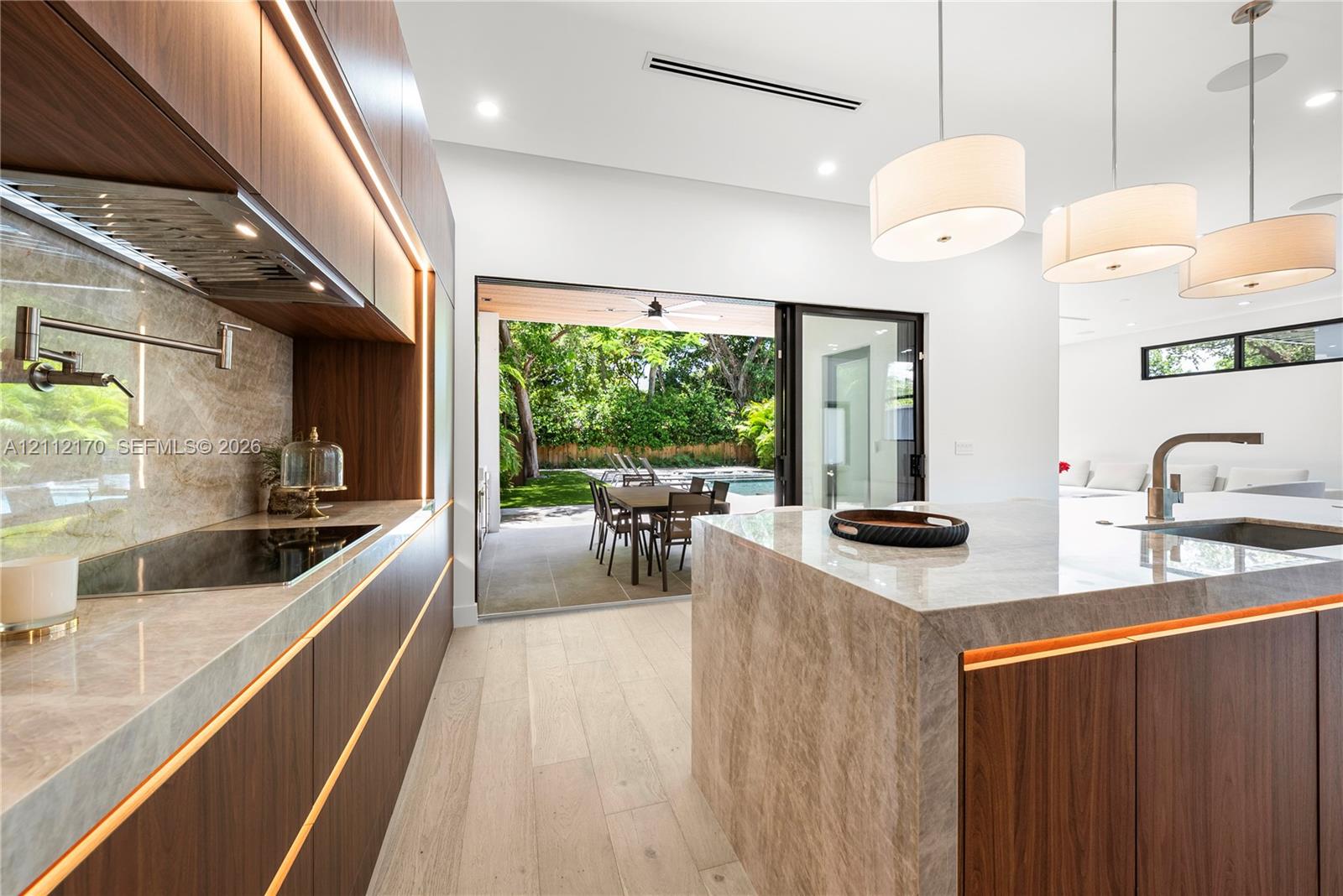 Kitchen featuring flat-panel wood cabinetry with integrated under-cabinet lighting, stone waterfall-edge island, wood-finish flooring, and wall-mounted pot filler faucet