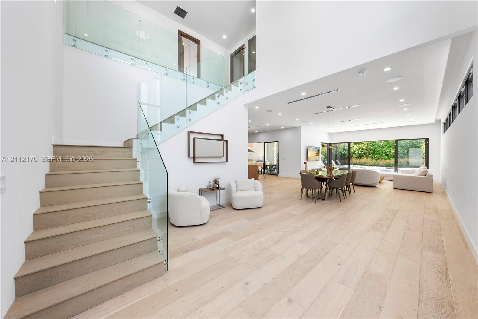 Open-concept interior featuring light wood-finish flooring, staircase with glass railings, recessed lighting, and expansive floor-to-ceiling glass sliding doors