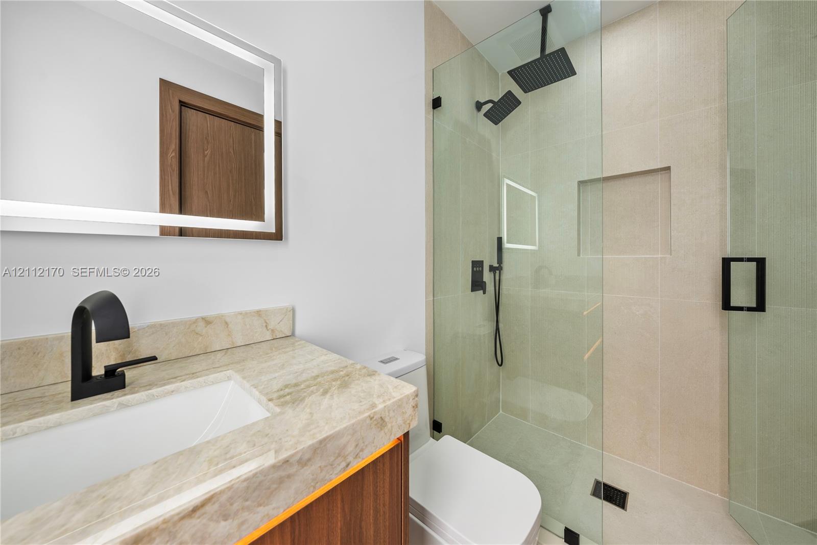 Bathroom featuring a wood-finish vanity with a stone countertop, an integrated rectangular sink, a frameless glass walk-in shower with matte black dual shower heads, and an LED-backlit mirror
