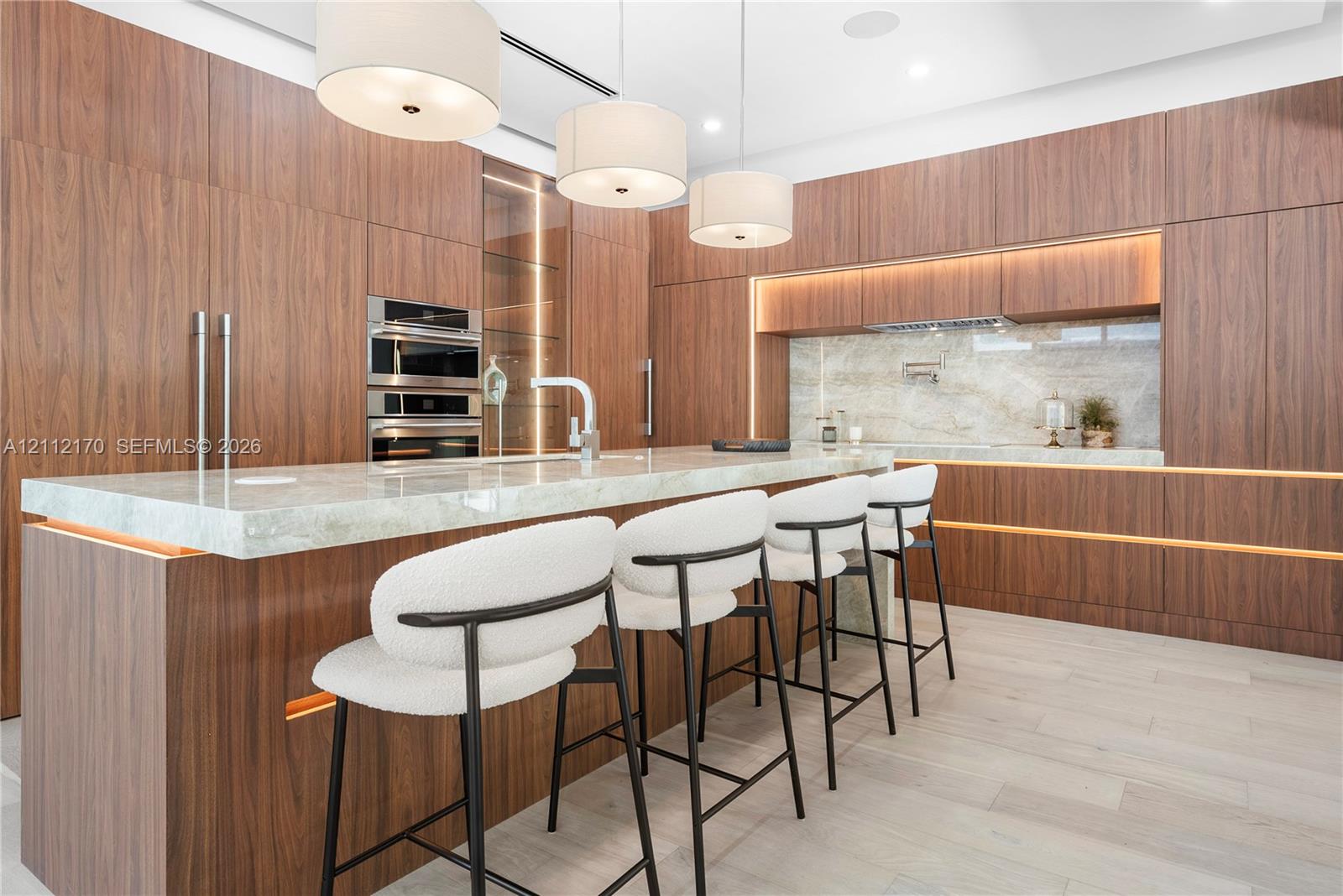 Custom wood-finish cabinetry, stone countertop island, built-in double ovens, natural stone backsplash with pot filler, and recessed under-cabinet lighting