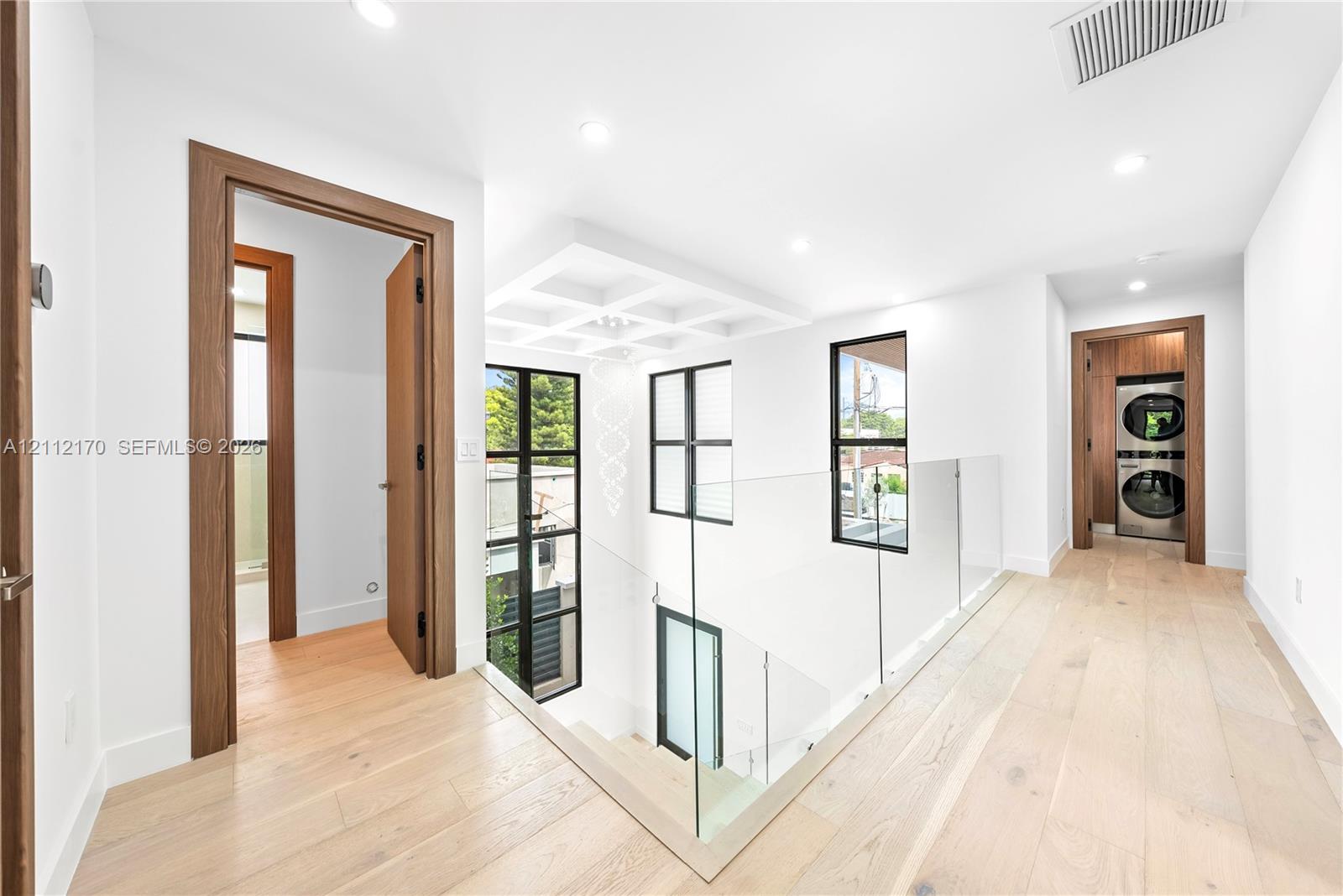 Hallway featuring light-toned wood-finish flooring, glass panel railing, coffered ceiling detail, and integrated recessed lighting