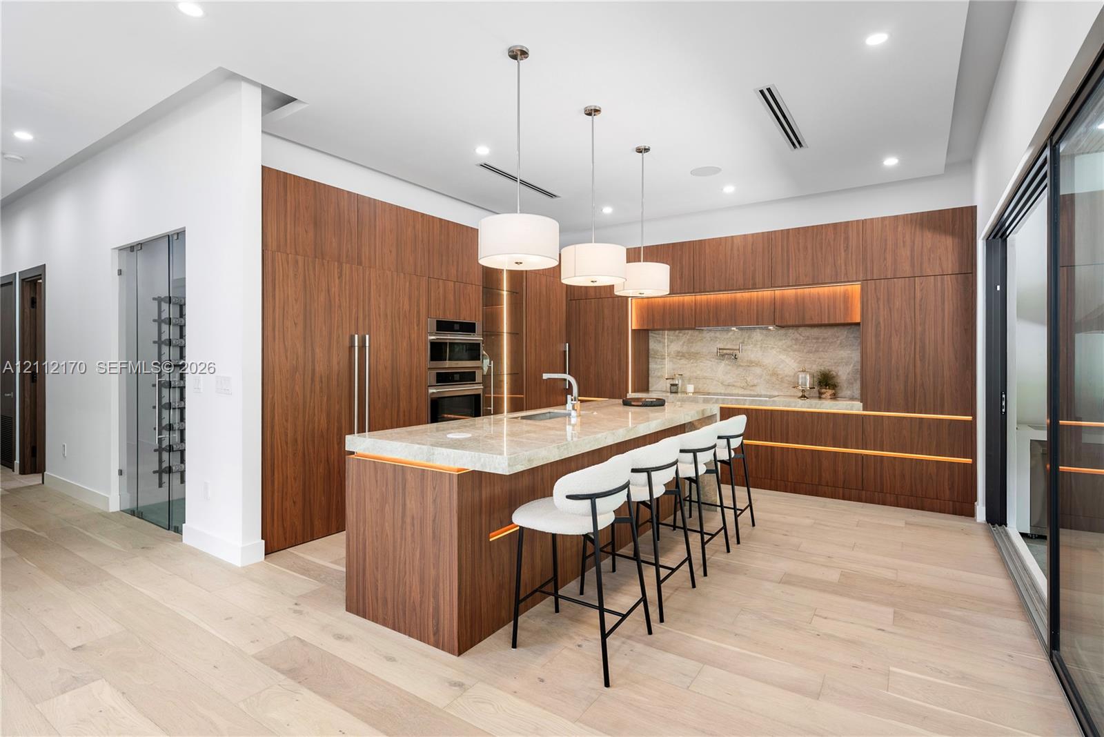 Modern kitchen featuring flat-panel wood cabinetry, a waterfall-edge stone island, built-in stainless steel double ovens, recessed lighting, and a glass-enclosed wine storage cabinet