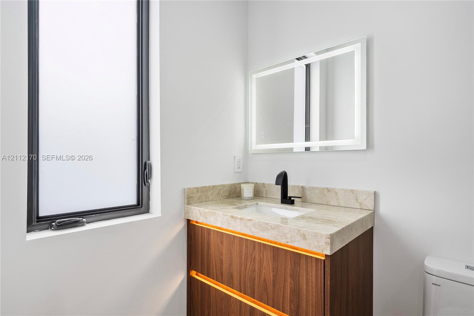 Modern vanity with wood-finish cabinetry, stone countertop, integrated undermount sink, matte black faucet, and rectangular LED-backlit mirror