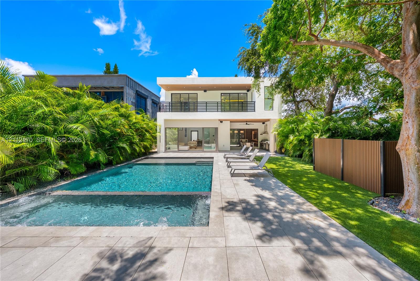 Modern two-story residence featuring a private pool with integrated spa, large-format tile patio, upper-level balcony with horizontal railing, and manicured green space