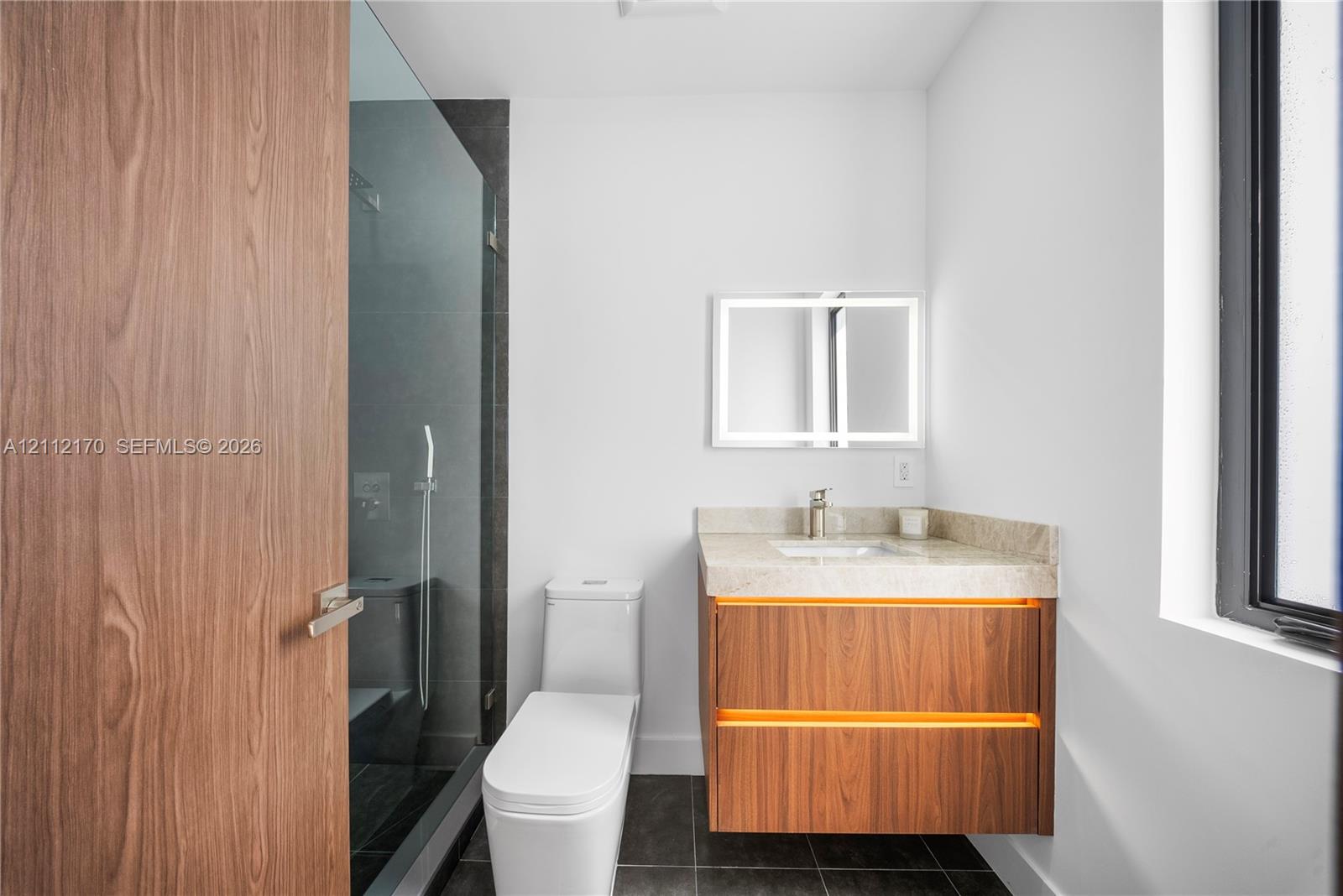 Floating wood-finish vanity with stone countertop, integrated LED lighting, glass-enclosed shower, and rectangular LED-backlit mirror