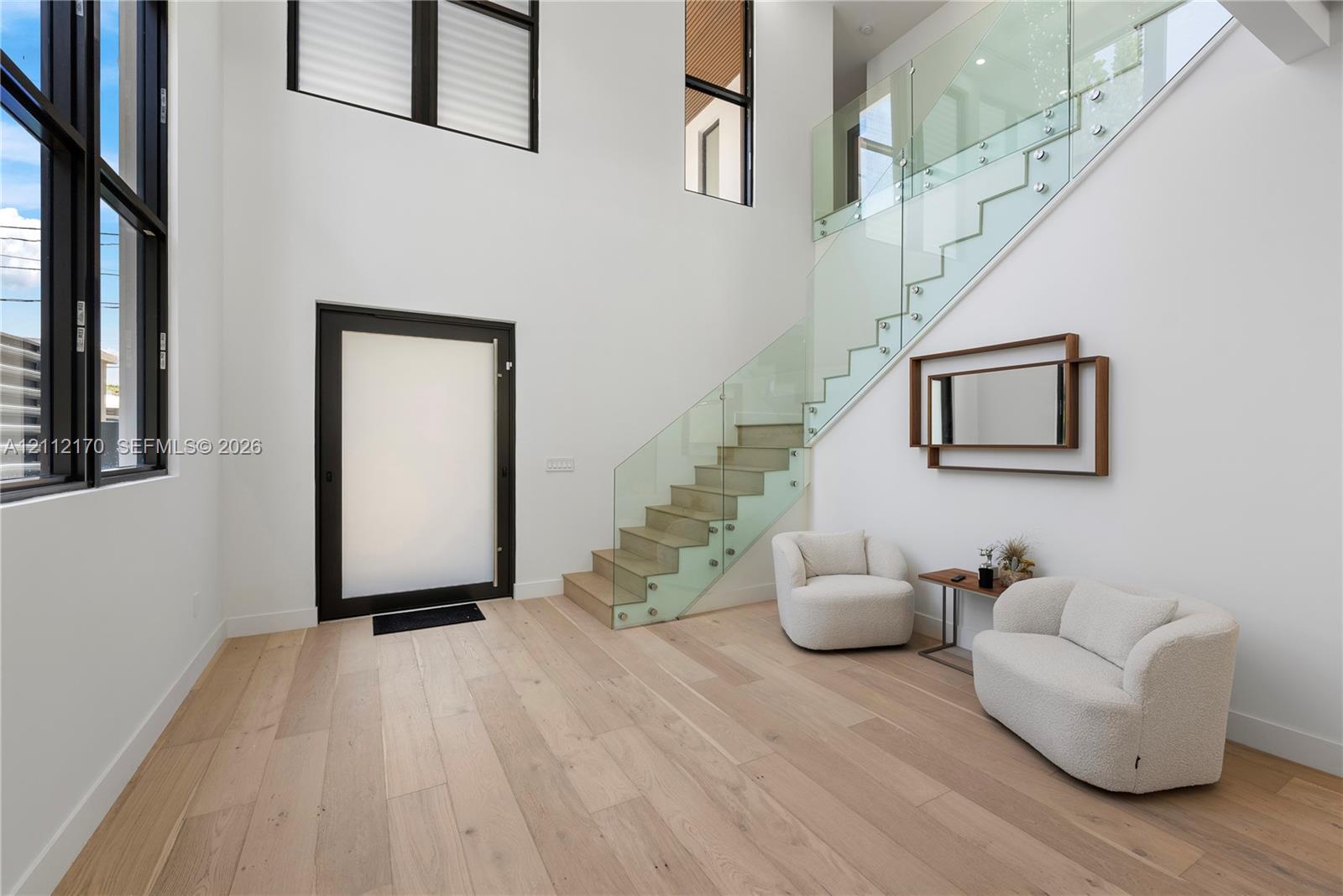 Double-height entry featuring light-toned wood-finish flooring, floor-to-ceiling windows, and a staircase with glass panels and metal hardware