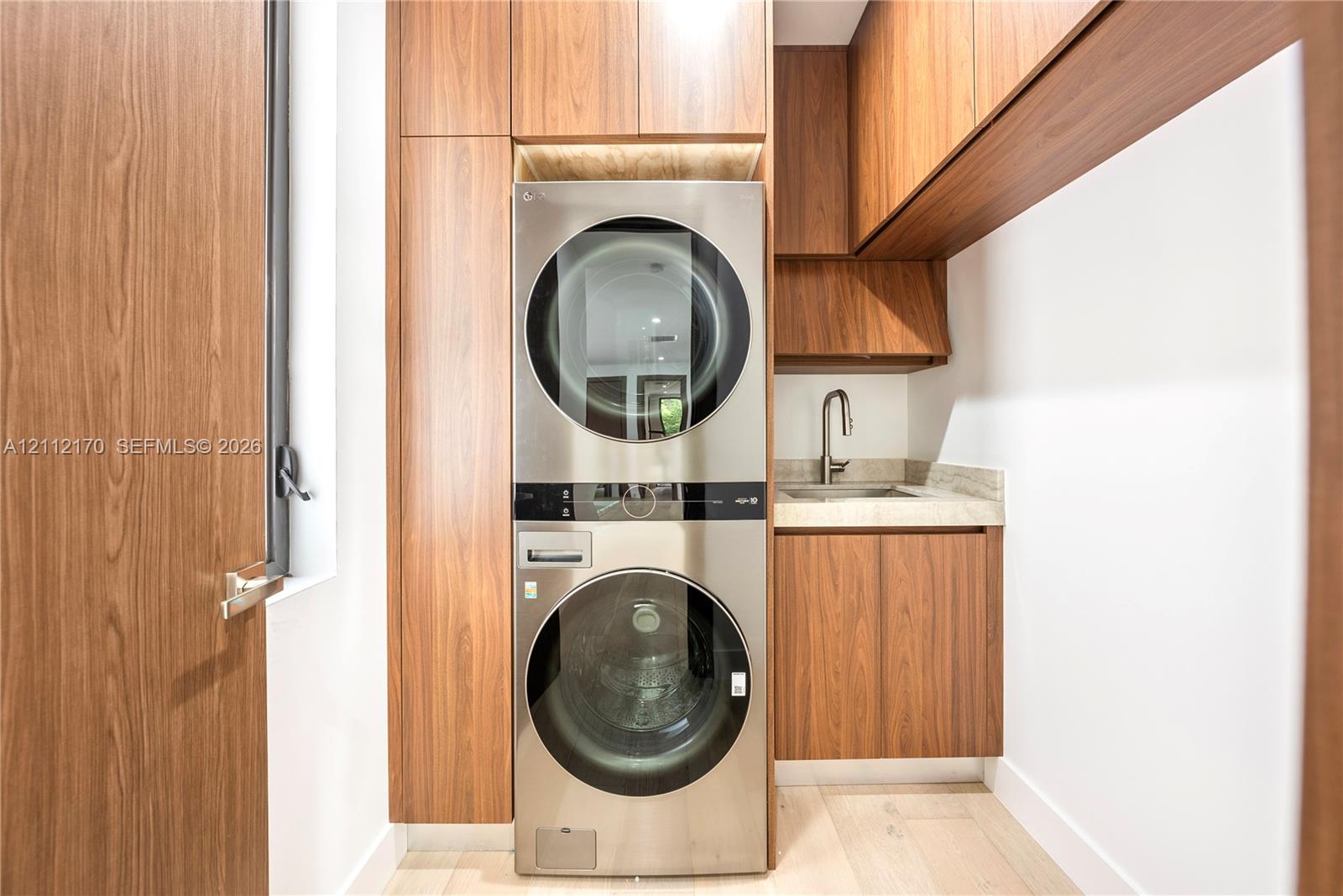 Utility room featuring custom wood-finish cabinetry, stainless steel stacked laundry appliances, integrated sink, and light-toned countertop