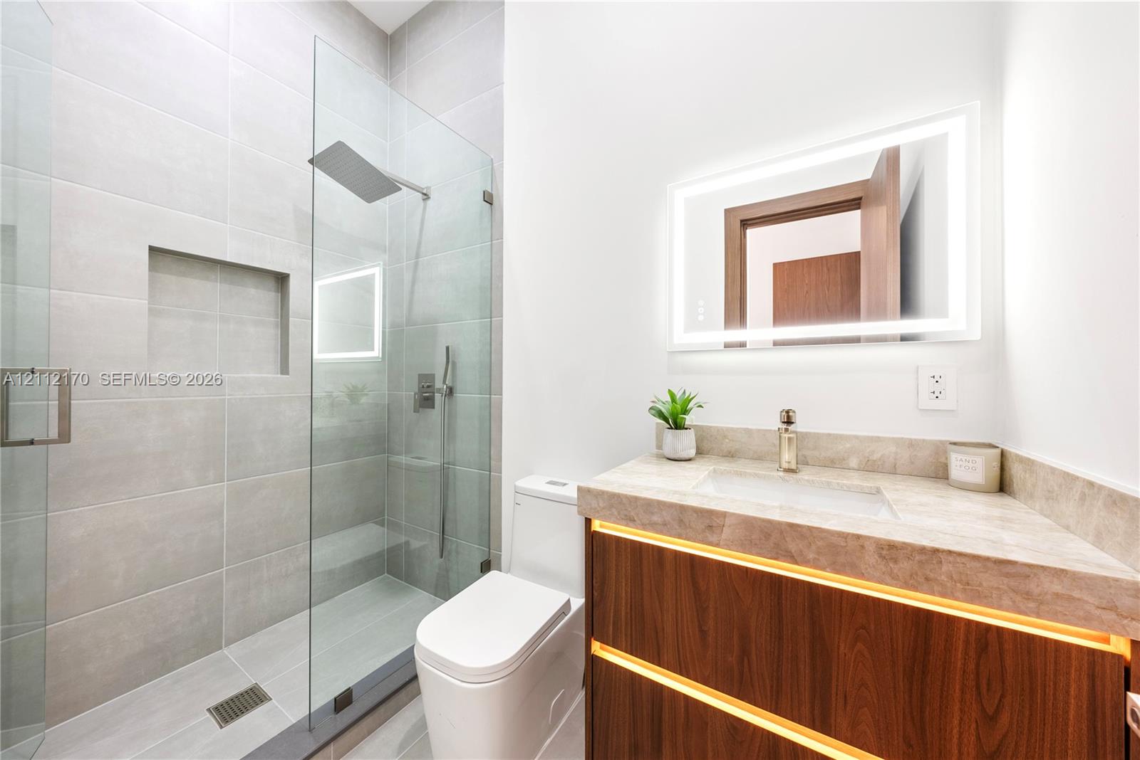 Floating wood-finish vanity with under-cabinet lighting, stone countertop with integrated sink, walk-in shower with large-format tile and built-in recessed niche, frameless glass shower enclosure, and rectangular LED-backlit mirror