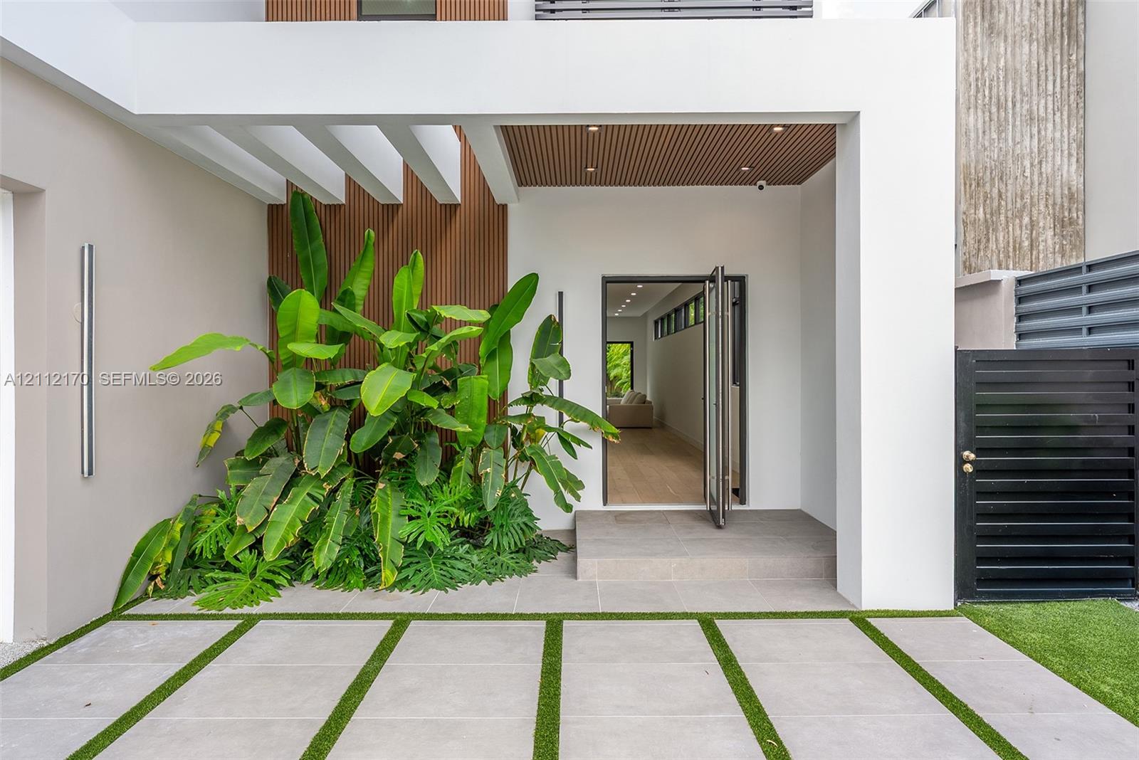 Modern entryway featuring a recessed porch with vertical wood-slat accent paneling, a concrete tile walkway with integrated turf borders, and a large-format tile porch landing