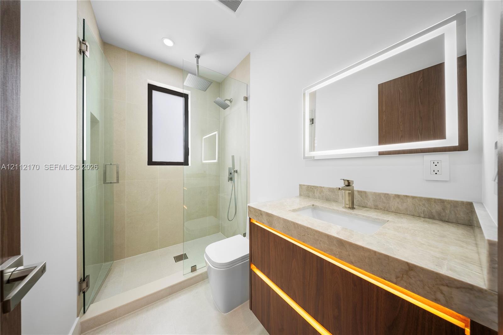 Floating vanity with integrated under-cabinet lighting, stone-top counter, walk-in shower with glass enclosure, rain-style showerhead, and wall-mounted LED-backlit mirror