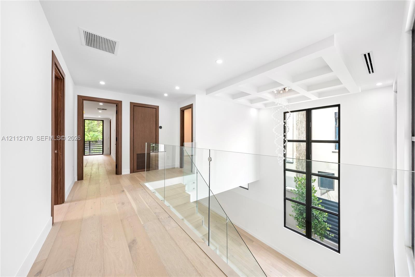 Hallway featuring wood-finish flooring, coffered ceiling detail, frameless glass stair railing, and metal-framed windows
