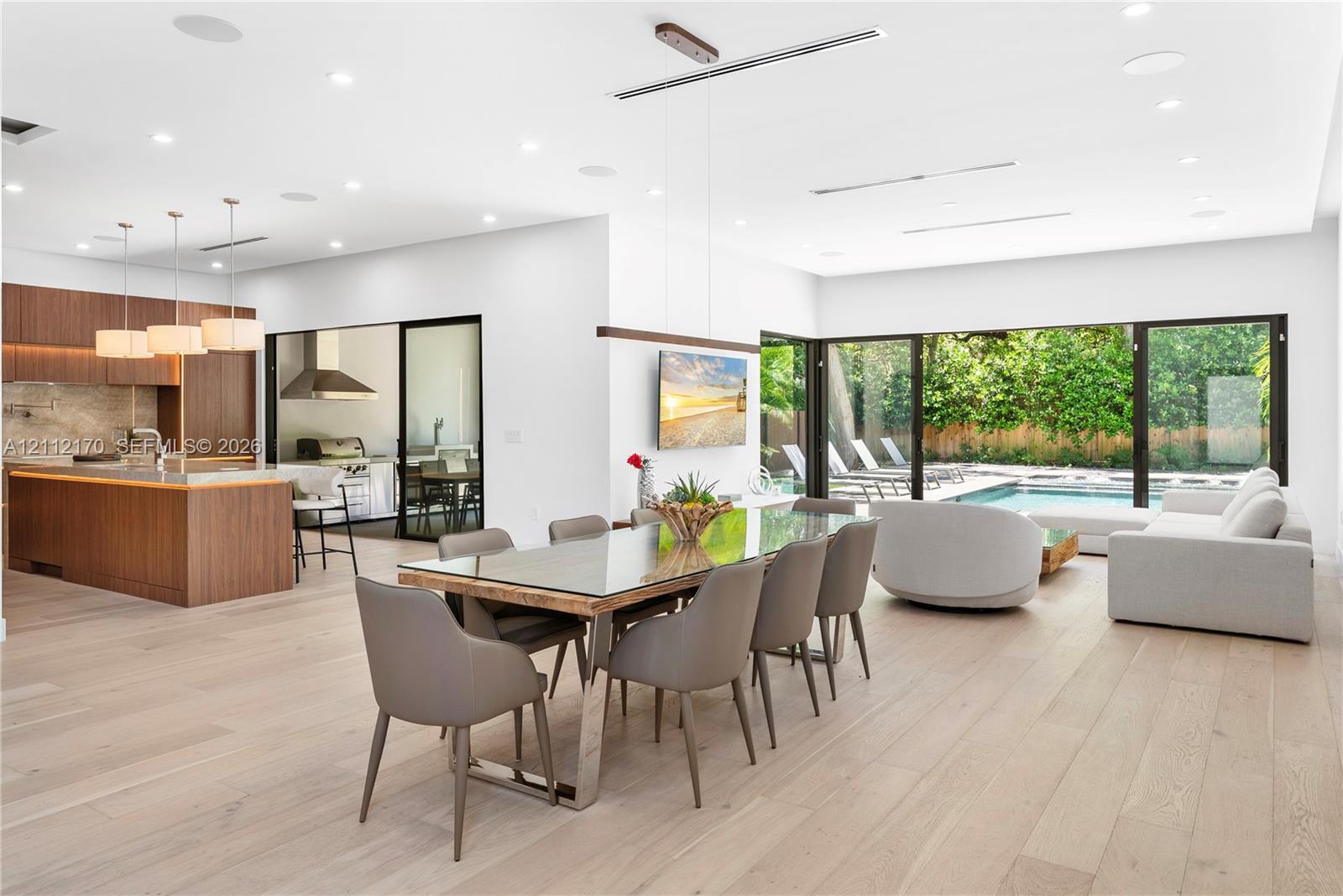 Open-concept interior featuring light wood-finish flooring, a kitchen island with stone countertop, flat-panel wood cabinetry, recessed lighting, and expansive floor-to-ceiling glass sliding doors