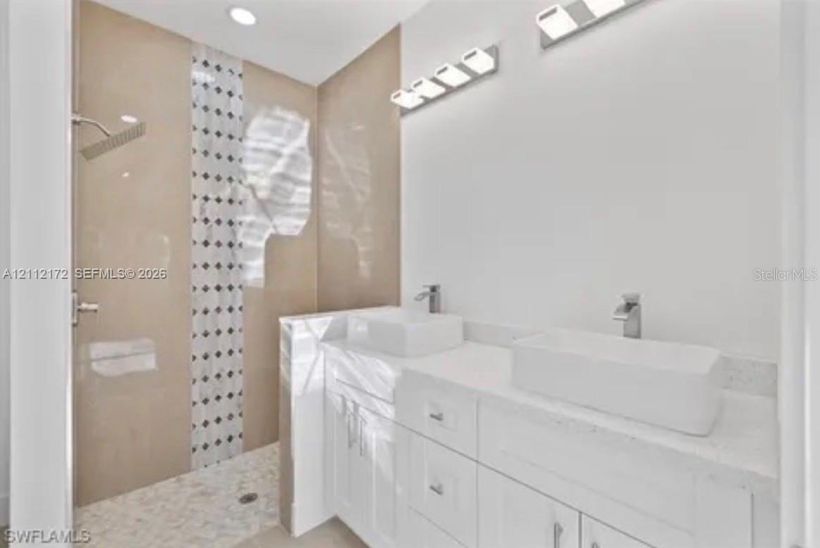 Dual white vessel sinks with chrome faucets, stone-topped vanity cabinetry, and a walk-in shower featuring neutral-toned tile work with a vertical mosaic accent