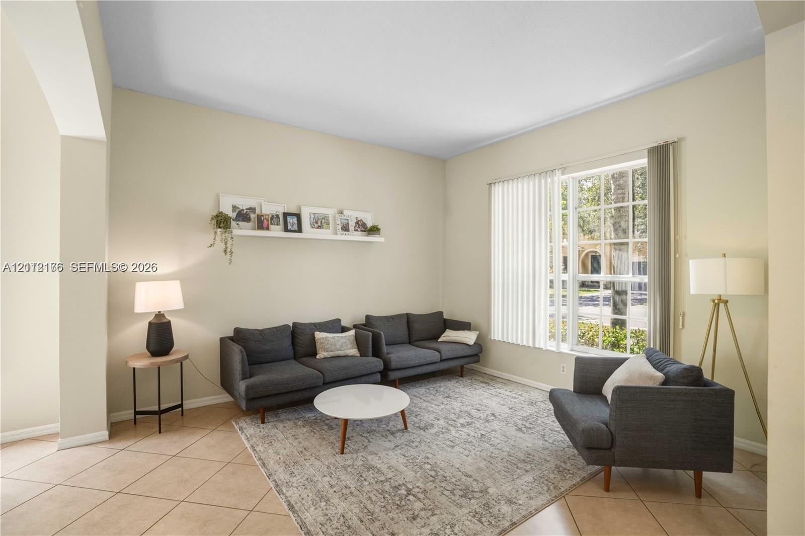 Living area featuring high ceilings, neutral wall tones, tile flooring, and a large multi-pane window with vertical blinds Living area featuring high ceilings, neutral wall tones, tile flooring, and a large multi-pane window with vertical blinds