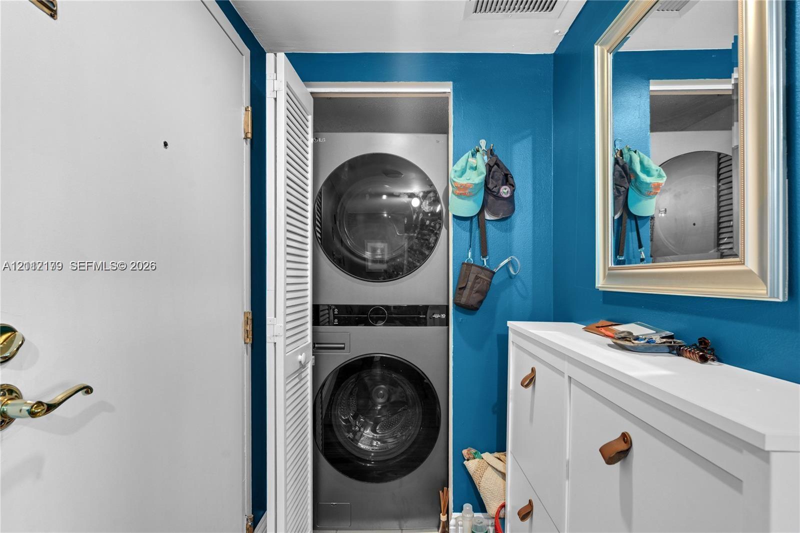 Stackable stainless steel washer and dryer unit, wall-mounted mirror with a metallic frame, and white louvered closet doors