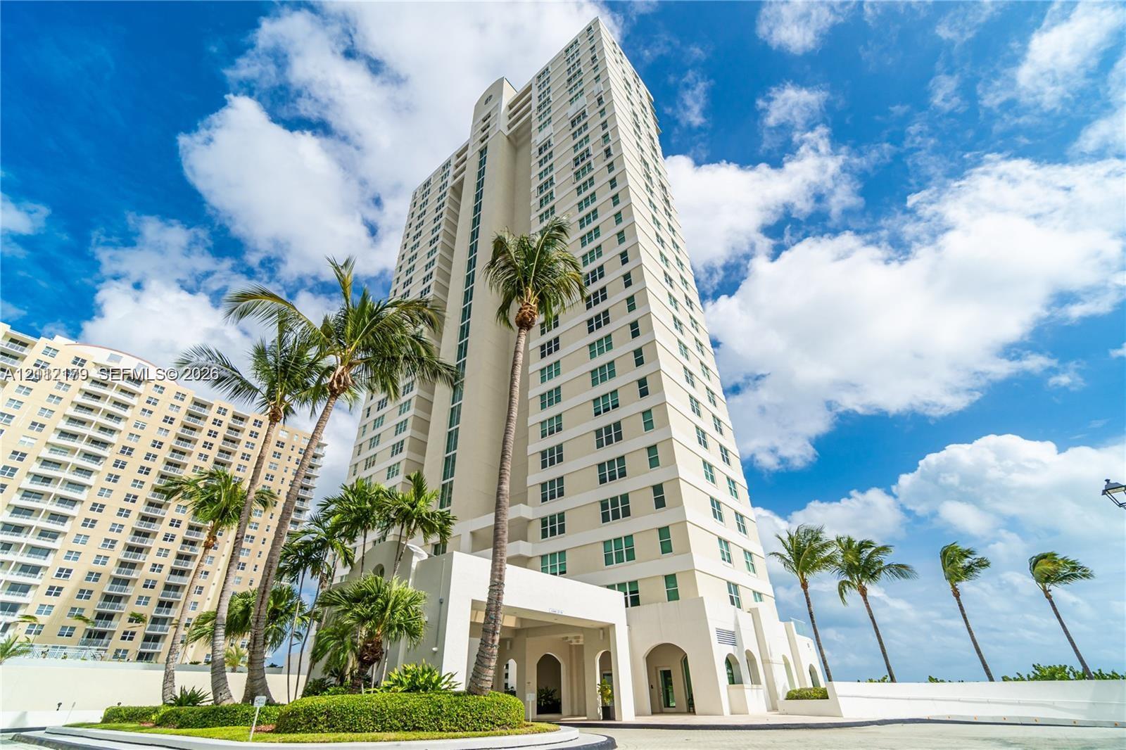 High-rise residential tower featuring a light-toned stucco facade, arched ground-level entryways, and manicured tropical landscaping with mature palm trees