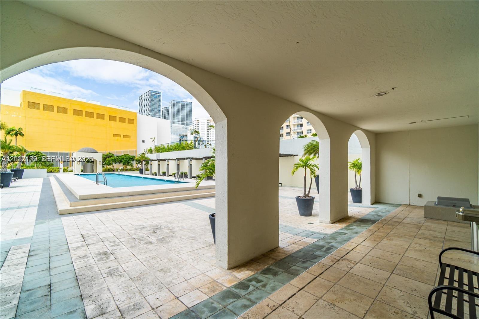 Covered patio featuring arched architectural details and stone-tile flooring, overlooking a swimming pool with tiered concrete decking