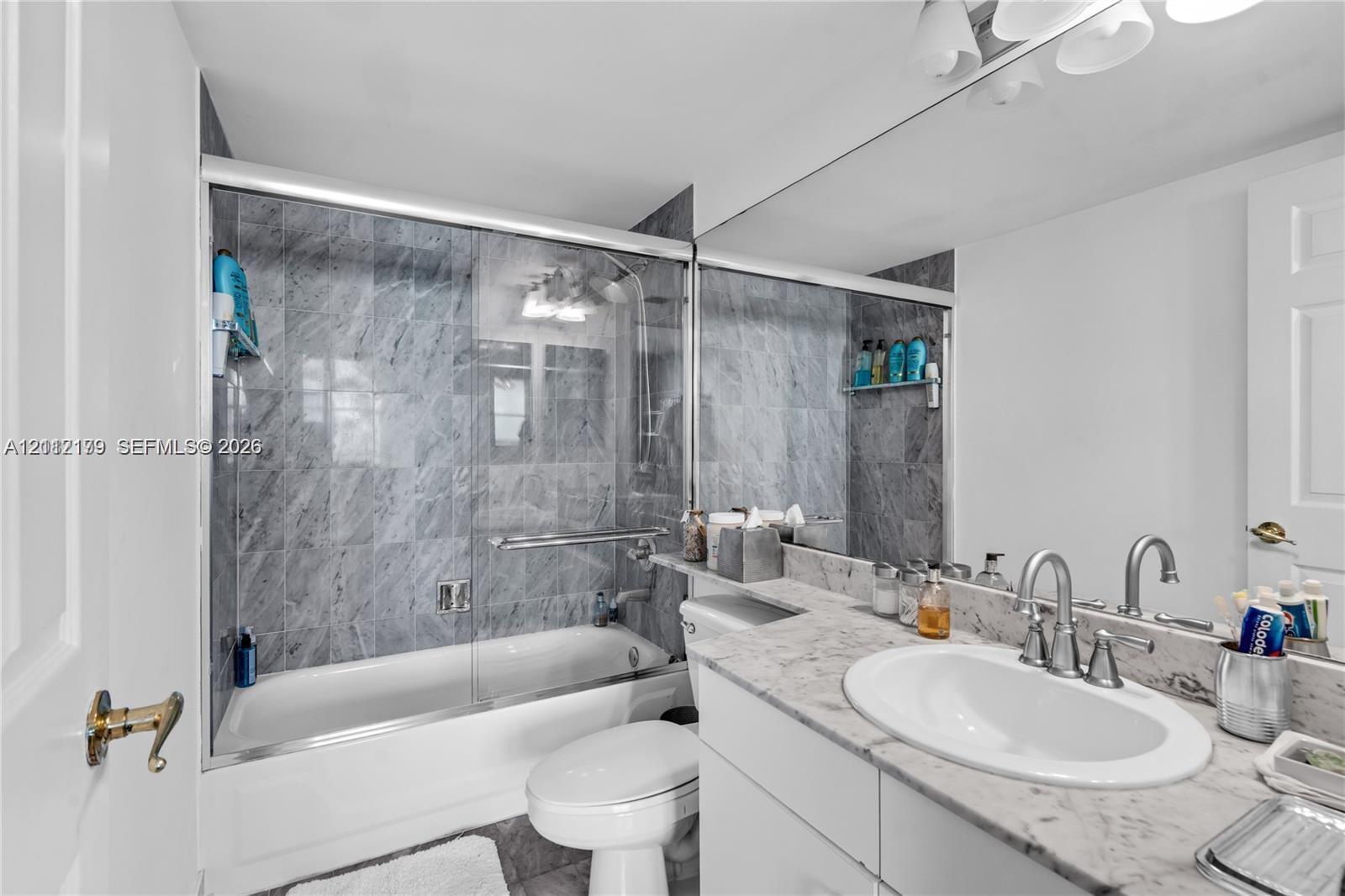 Full-width vanity with stone countertop and undermount sink, shower-tub combination with glass enclosure, and floor-to-ceiling stone tile surround