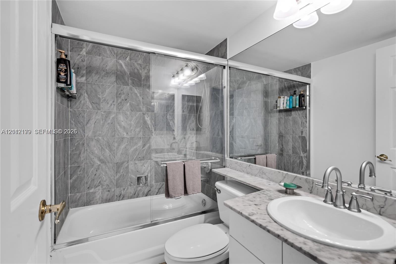 Bathroom featuring a shower-tub combination with glass enclosure, full-height stone-finish wall tile, a stone-top vanity with integrated sink, and an expansive wall-to-wall vanity mirror