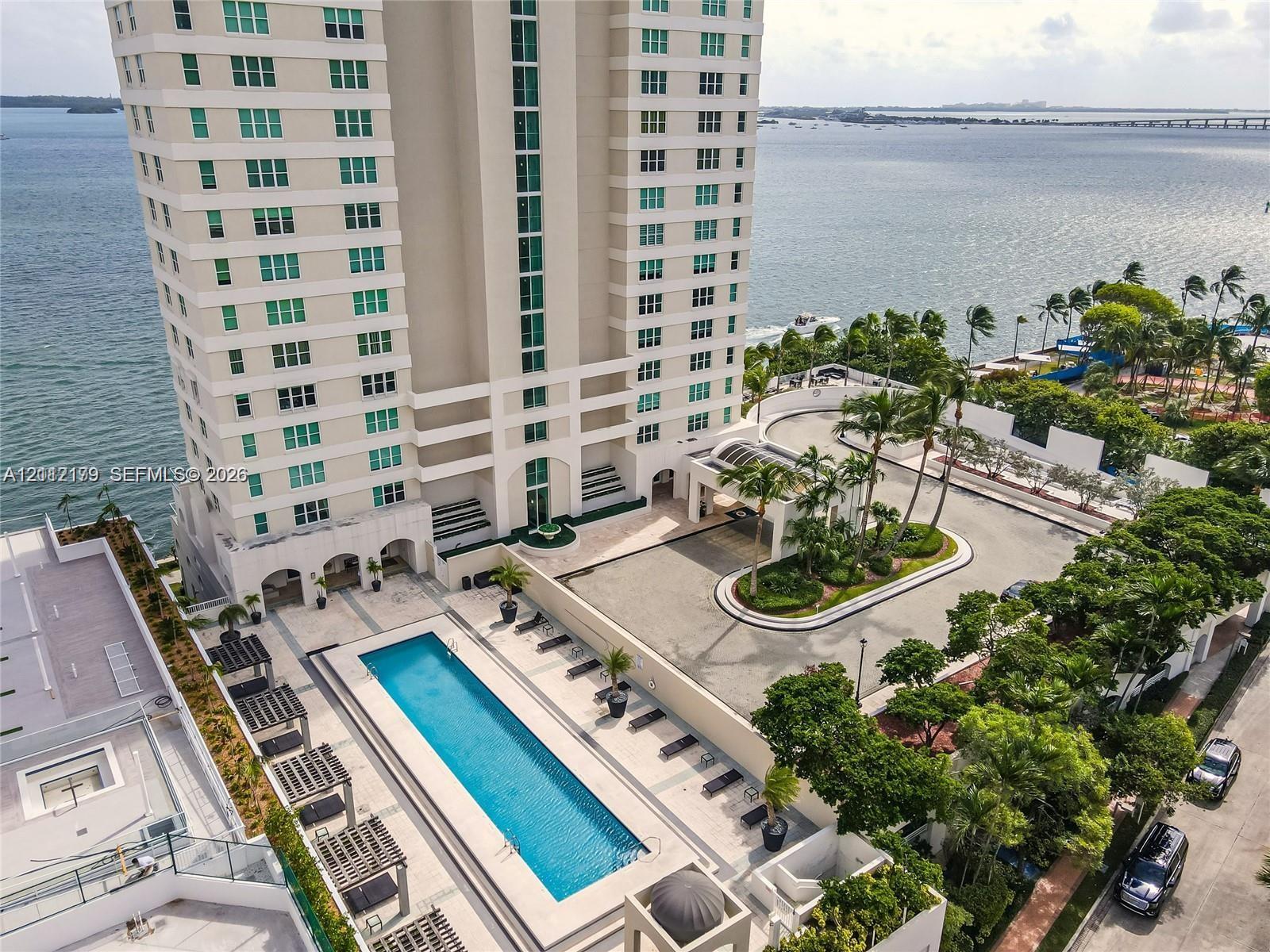 High-rise residential building featuring an expansive outdoor swimming pool, paved pool deck with shade structures, porte-cochre entry, and surrounding mature landscaping, positioned on a waterfront site