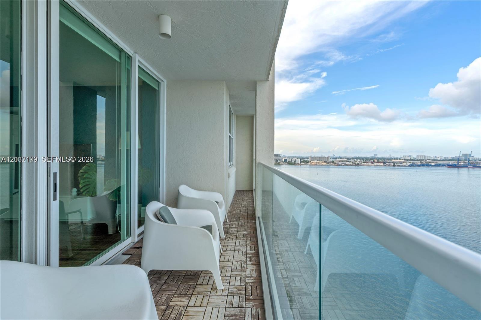Covered balcony featuring wood-finish deck tiling, glass panel railing, floor-to-ceiling sliding glass doors, and direct water frontage