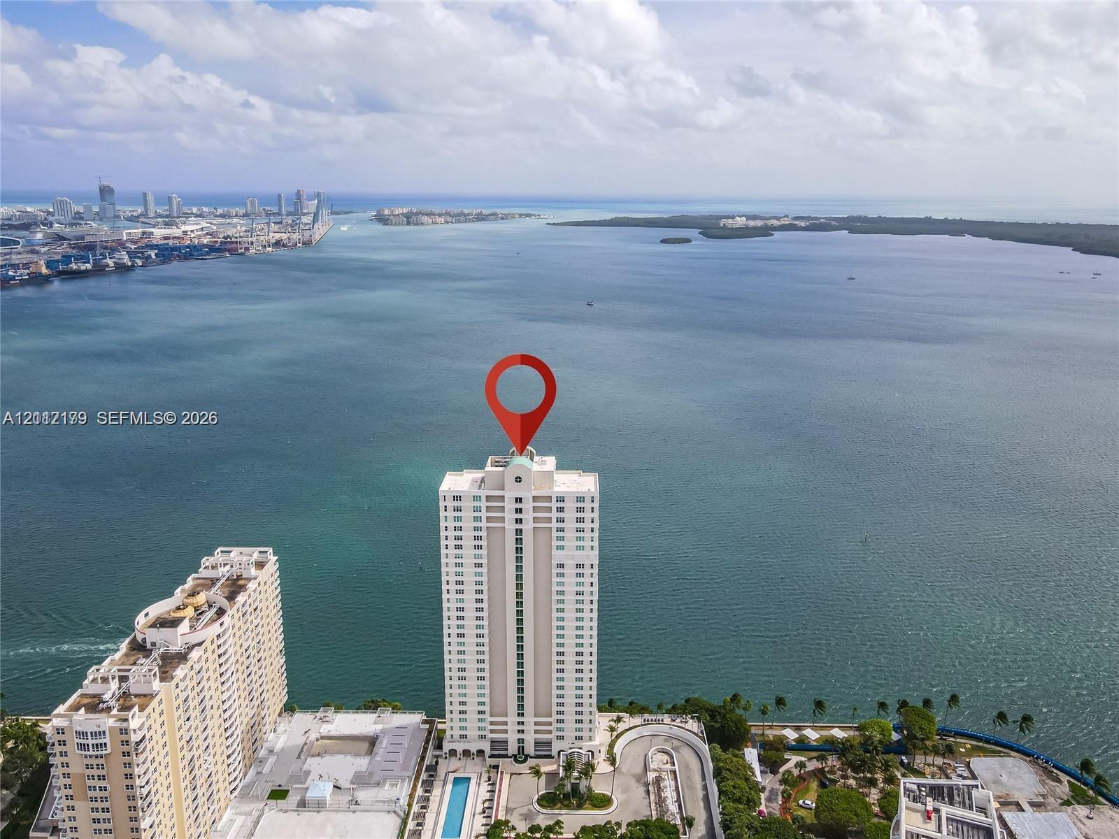 Aerial perspective of a high-rise waterfront building featuring an outdoor swimming pool, paved surface parking, and extensive shoreline exposure