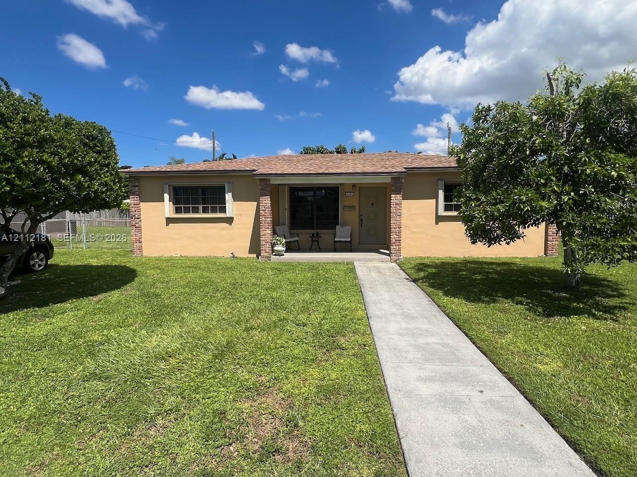 Single-story residence featuring a tan stucco exterior, brick column accents, a covered front entryway, and a concrete walkway set amidst a level lawn