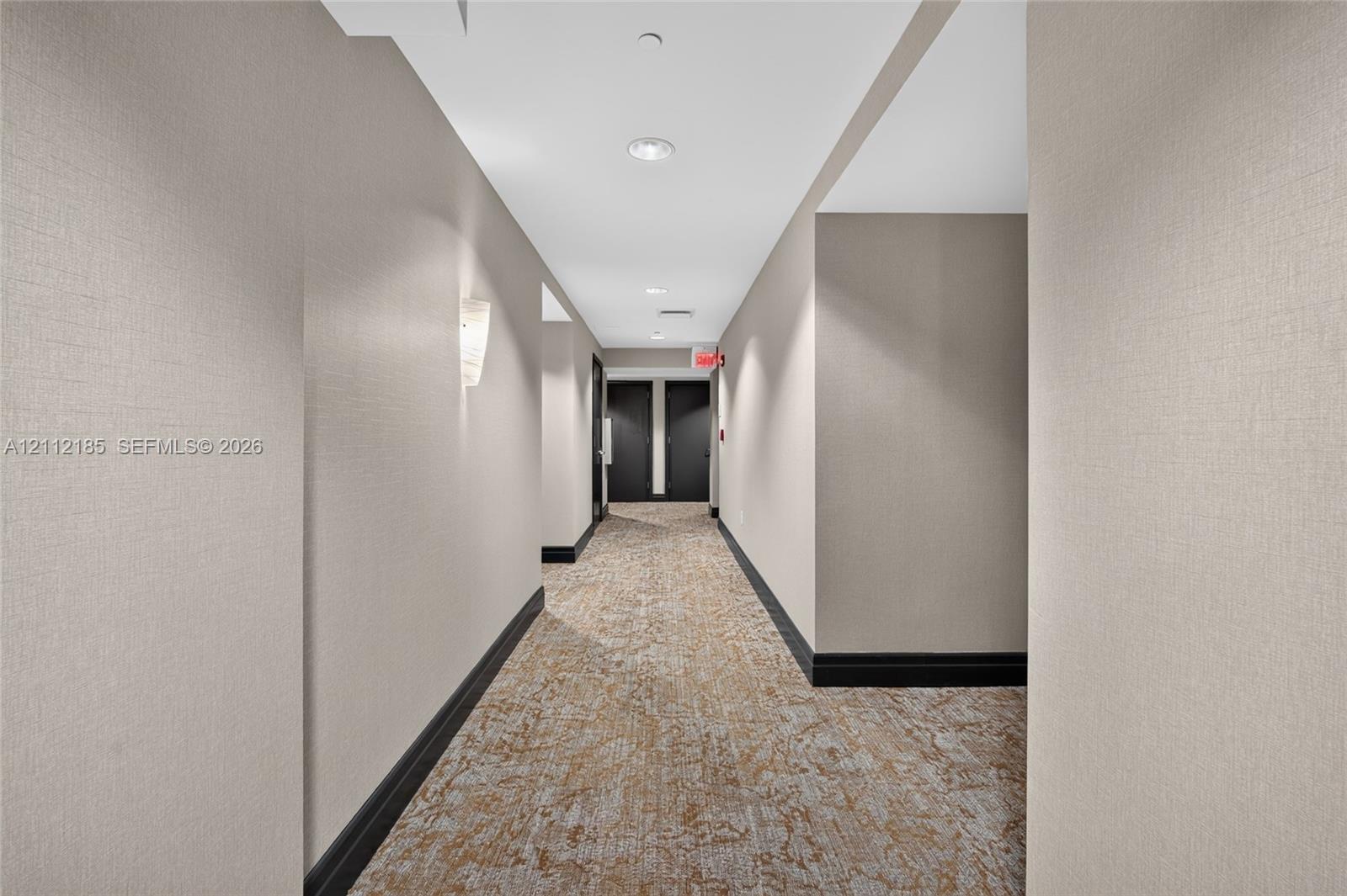 Hallway featuring textured neutral wall covering, patterned carpeted flooring, contrasting dark trim, and wall-mounted sconce lighting