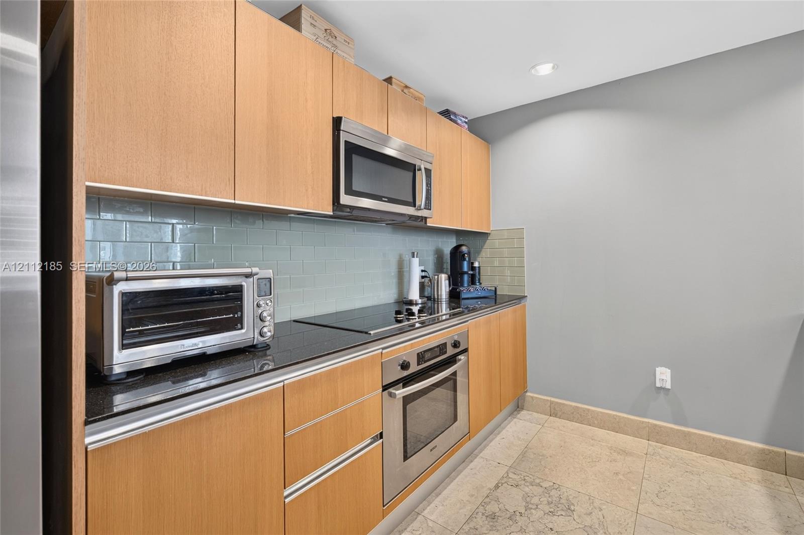 Kitchen featuring flat-panel wood cabinetry, black countertops, a subway tile backsplash, stainless steel appliances, and tile flooring