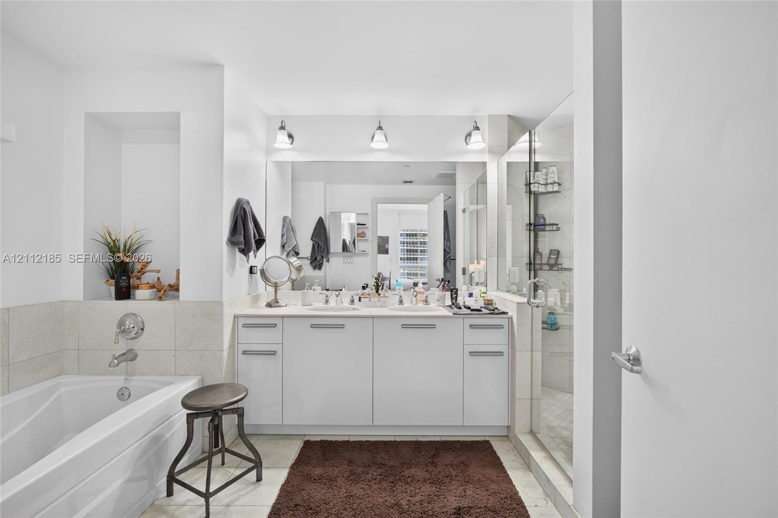Bathroom featuring dual-sink vanity with light-toned cabinetry, built-in recessed wall shelving, soaking tub with wall-mounted chrome fixtures, and glass-enclosed shower with tiled surround