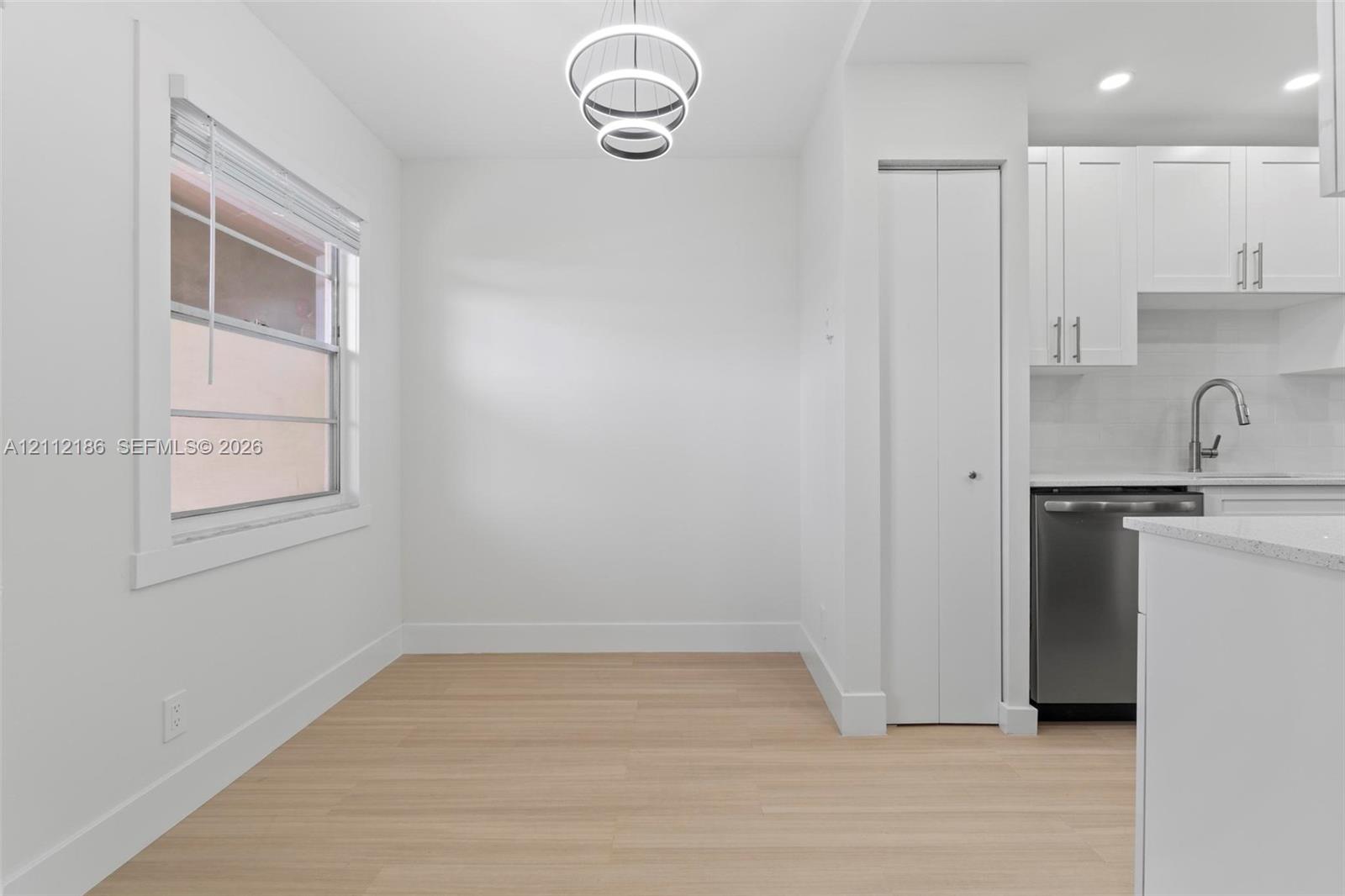 Light-filled area featuring wood-finish flooring, a modern three-ring pendant light, a recessed folding-door closet, and white cabinetry with a stainless steel dishwasher
