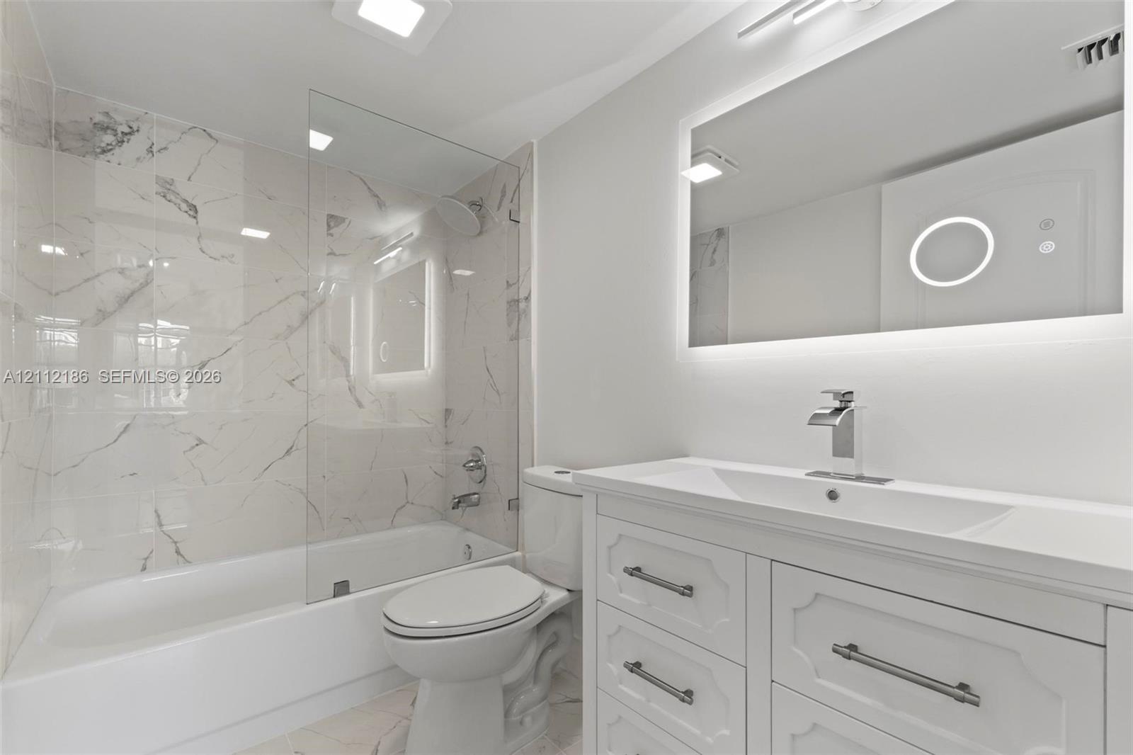 Bathroom featuring white custom cabinetry with brushed hardware, stone-finish wall tile surround, glass panel shower enclosure, and integrated LED-backlit mirror