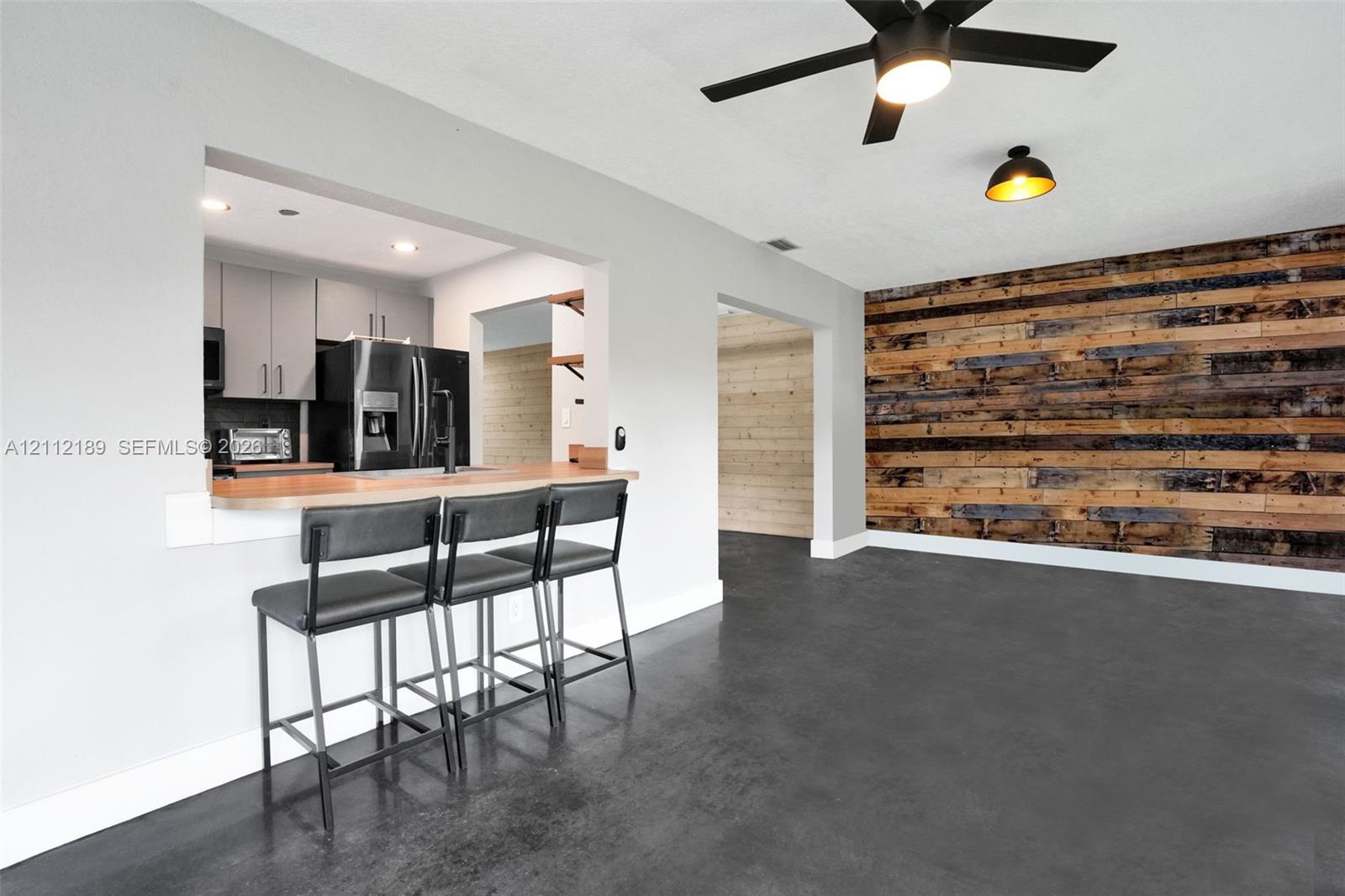 Interior space featuring a dark polished concrete floor, a horizontal wood-plank accent wall, a kitchen pass-through with a breakfast bar, and a ceiling-mounted matte black fan with integrated lighting