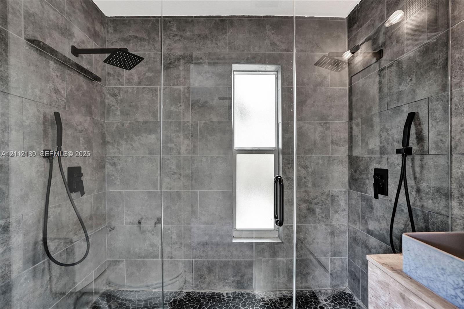 Walk-in shower featuring large-format grey stone wall tile, black matte rainfall showerheads, handheld shower fixtures, a glass door enclosure, and a stone-tiled floor