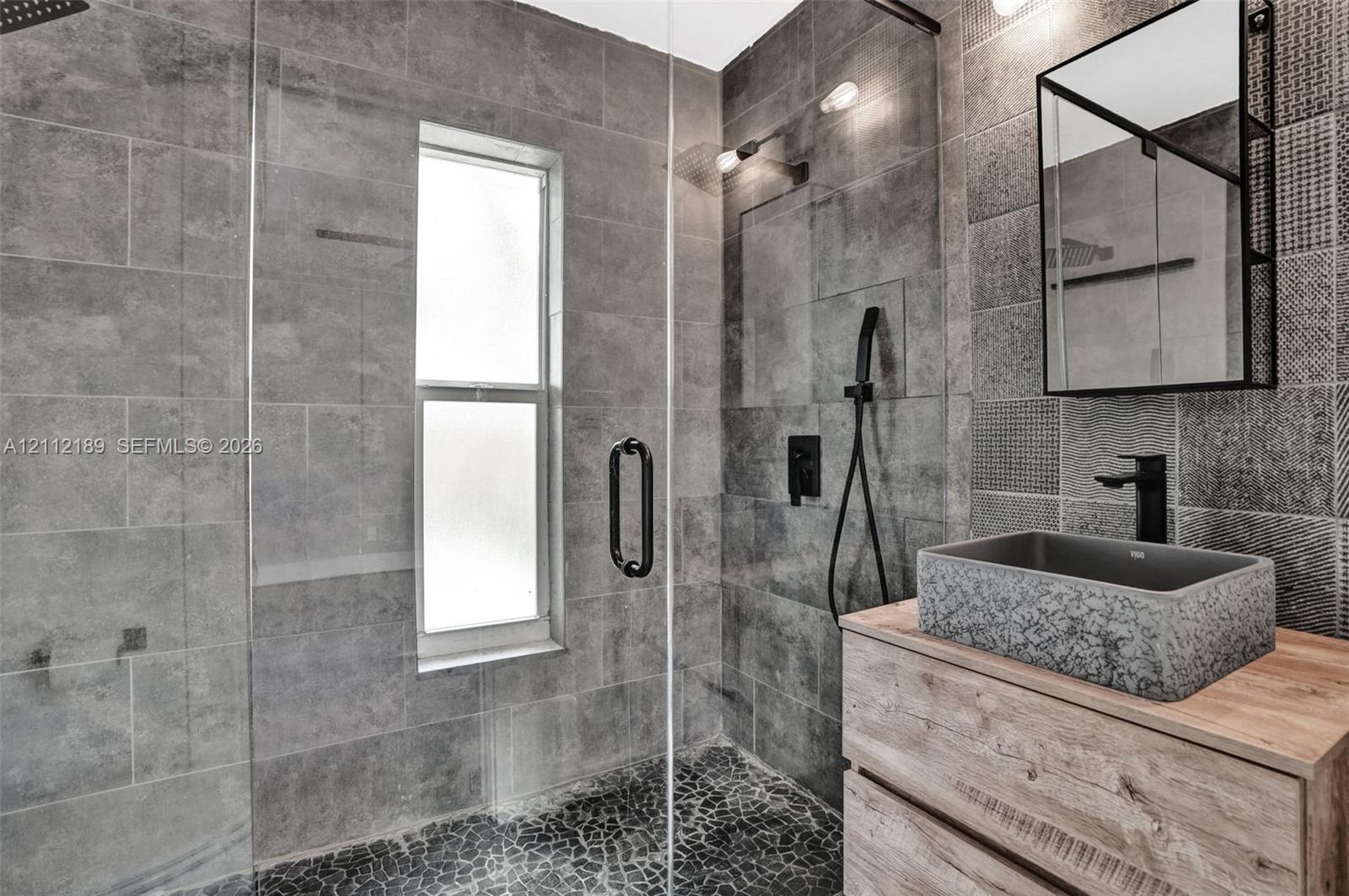 Bathroom featuring a walk-in shower with floor-to-ceiling textured tile, a frameless glass enclosure, pebble-stone flooring, a wood-finish vanity with a stone vessel sink, and a framed mirror