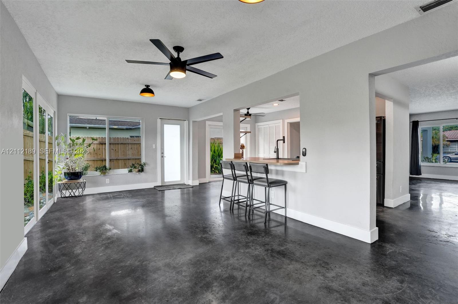 Polished concrete flooring, pass-through kitchen window with bar seating, matte black ceiling fan, and large windows with natural light exposure